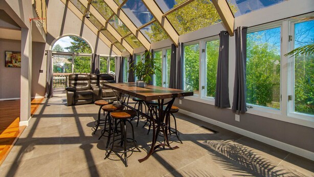 A sunroom with a glass ceiling, a dining table with stools, leather sofas, and large windows offering a view of greenery.