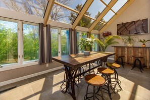 Ein Wintergarten mit Holztisch, Barhocker, großen Fenstern und Topfpflanzen.