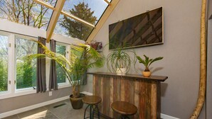 A room with a wooden bar, two stools, a large window, and potted plants.