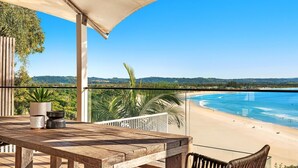 Ein Holztisch mit einem Topfpflanze und einer Schüssel, mit Blick auf einen Strand mit klarem, blauem Wasser und einer entfernten bewaldeten Gegend.