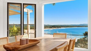 Ein Essbereich mit einem Holztisch und Stühlen, einem großen Fenster mit Blick auf Strand und Meer und einer Schiebetür zum Außengelände.