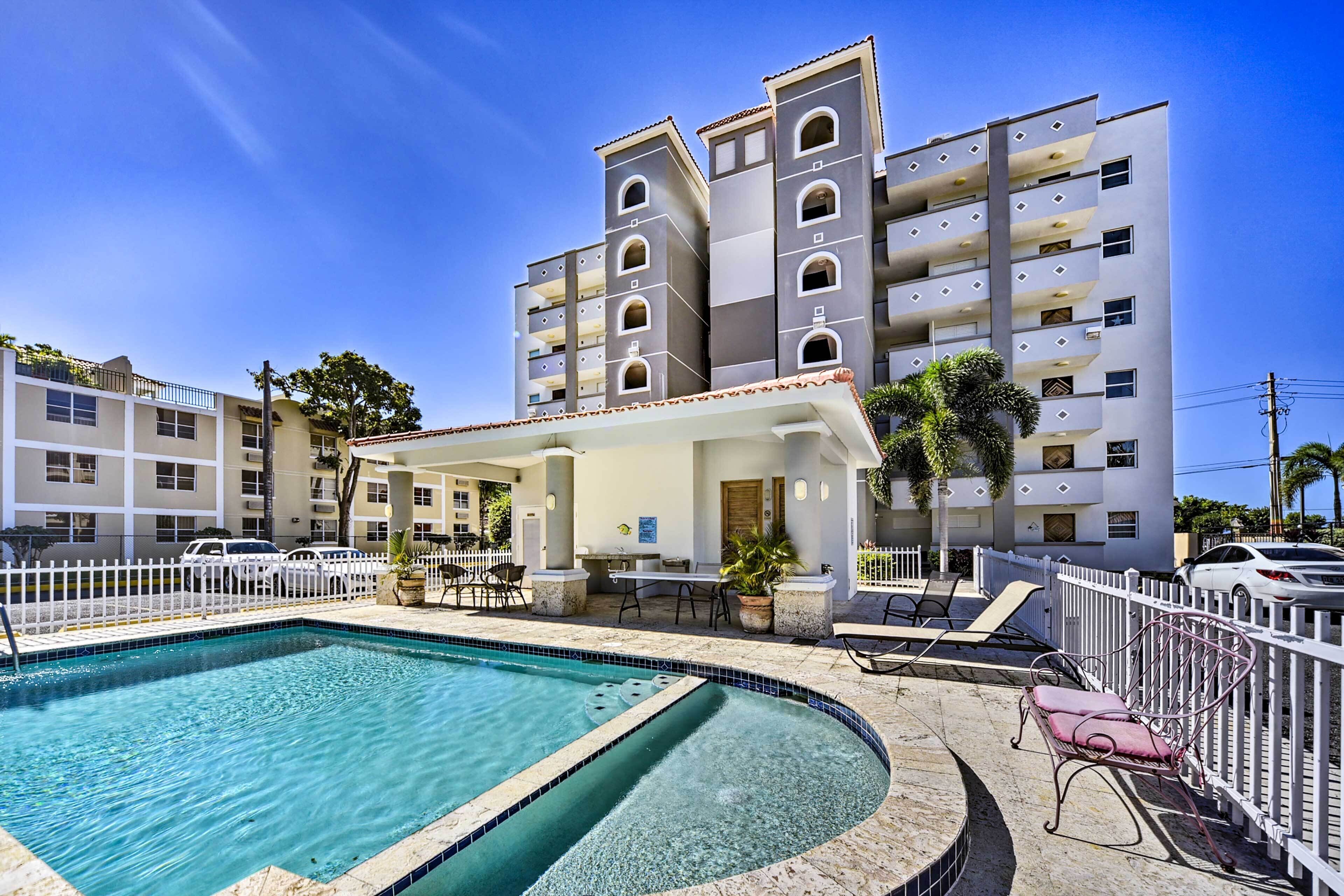 A hotel with a swimming pool, outdoor seating, and a white fence.