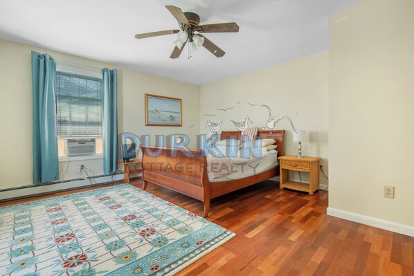 A bedroom with a wooden bed, a ceiling fan, a window with curtains, a wall-mounted painting, and a patterned rug.