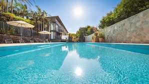 A clear blue pool with lounge chairs and umbrellas, surrounded by stone walls and tropical plants.
