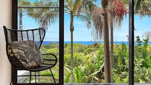 A black wicker chair with a patterned cushion in a room with a large window overlooking a tropical landscape with palm trees and a view of the ocean.