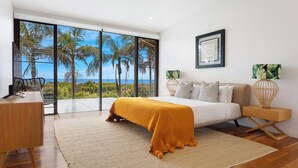 A bedroom with a bed, a desk, and a chair, a large window with a view of palm trees and the ocean, and a framed artwork on the wall.