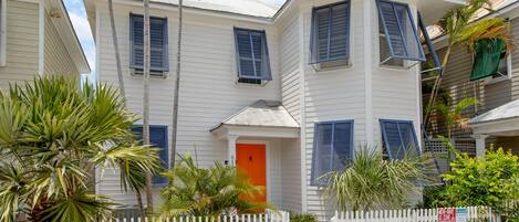 A white two-story house with blue shutters, a white picket fence, and palm trees.