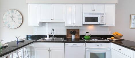 A kitchen with white cabinets, a black countertop, a white dishwasher, a white oven, a microwave, a sink, and a clock.