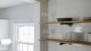 Een keuken met witte betegelde wanden, een houten plank met glazen en een raam met zonwering.