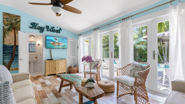 Ein geräumiges Wohnzimmer mit Holzboden, Deckenventilator, großem Fenster mit Vorhängen, Blick auf einen Pool und einem 'Hay West' Schild an der Wand.