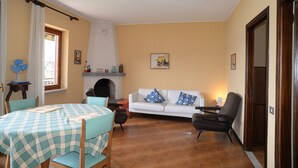 Un salon avec une cheminée, un canapé blanc orné de coussins bleus, une table ronde recouverte d’une nappe à carreaux et un ventilateur posé sur une table d’appoint.