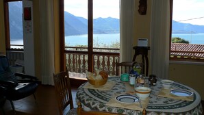 Un coin repas avec vue sur le lac et les montagnes, une table dressée avec des assiettes et un panier de pain, et une chaise agrémentée d’un coussin à motifs.