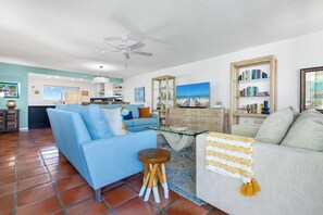 A living room with a blue sofa, a white sofa, a glass coffee table, and a ceiling fan.