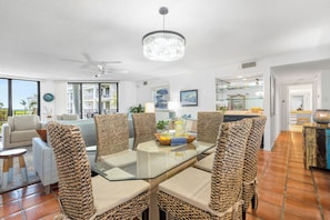 A modern living room with a glass-top dining table surrounded by woven chairs, a ceiling fan, and a chandelier.