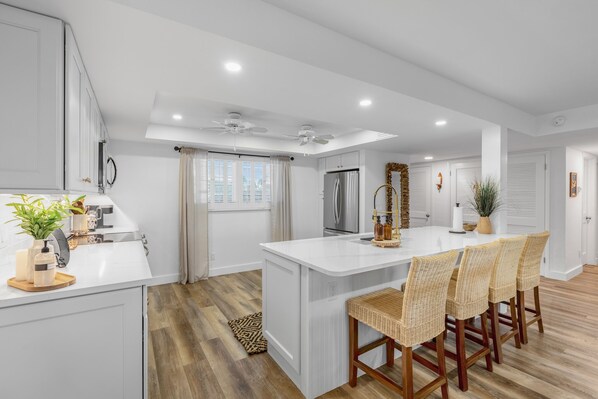 Cocina moderna con armarios blancos, una isla central con taburetes y un ventilador de techo, junto a un amplio ventanal con persianas.