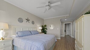 Une chambre à coucher comprenant un lit, des tables de chevet, un ventilateur de plafond, un miroir et un couloir menant à une porte.