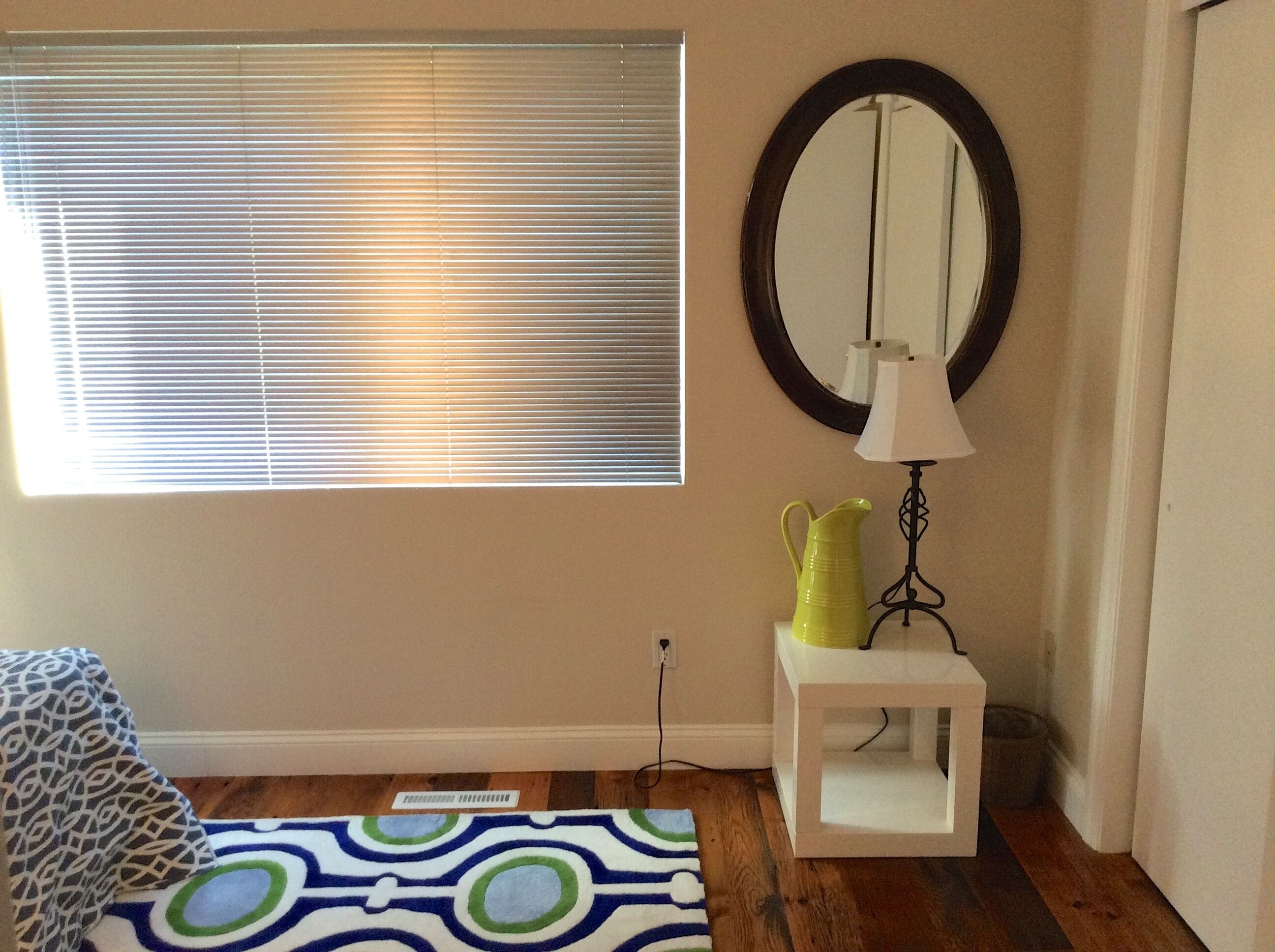 A room with a window with blinds, a mirror on the wall, a lamp on a small table, and a patterned rug on the floor.