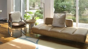 Een ruime woonkamer met een groot raam, een bank, een fauteuil en een tafeltje met een plant.