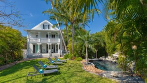 A large white house with a balcony, surrounded by a well-maintained lawn and palm trees. - SOUTHERNMOST BELLE~ Heated Pool, Gourmet Kitchen & Elegant Large Bedrooms! (Key West)