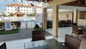 An outdoor patio area with wicker chairs and a glass-top table, colorful paper flags hanging from the ceiling, and a view of a building in the background. - Apartment in milionários beach, Ilheus - BA (Ilheus)