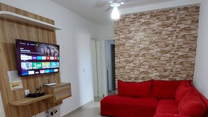 A living room with a flat-screen TV mounted on a wooden wall, a red sofa, and a small dog on the floor. - Apartment in milionários beach, Ilheus - BA (Ilheus)