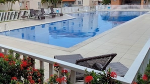 A swimming pool with a white deck, surrounded by a white fence and red flowers in the foreground. - Apartment in milionários beach, Ilheus - BA (Ilheus)