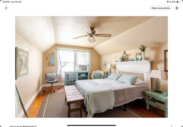 Une chambre à coucher comprenant un lit, un ventilateur de plafond, une fenêtre avec des rideaux, une chaise et une petite table avec une lampe.