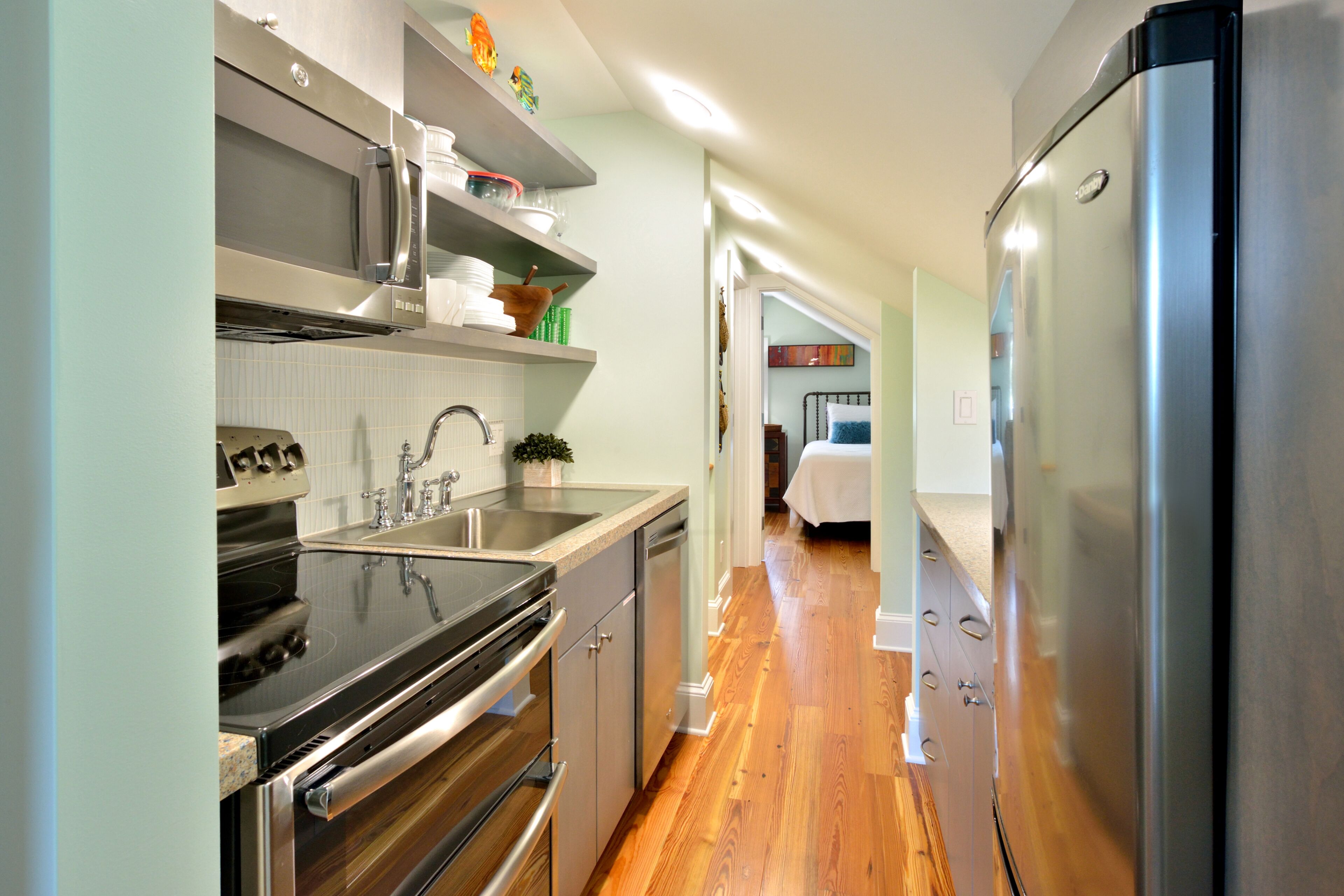 A modern kitchen with stainless steel appliances, wooden flooring, and a view into the adjacent bedroom.