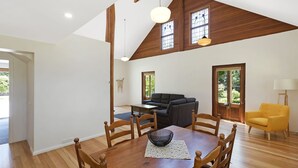Ein geräumiges Zimmer mit Esstisch, Stühlen, einer Couch und einem gelben Sessel. Zwei Fenster mit Holzrahmen und eine Decke mit Holzbalken sind vorhanden.