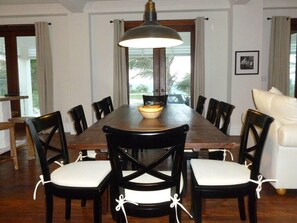 A dining room with a long wooden table, black chairs with white cushions, a large pendant light, and a view of the outdoors through large windows. - Private Beachfront Home w/Pool, Solar, Banks Rd, Walk to Restaurants (Governors Harbour)