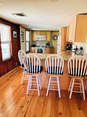 A kitchen with wooden cabinets, a microwave, a stove, and a clock on the wall. There are three white chairs with black cushions at a wooden island. - Cozy Home, Lots of Light, Three Blocks from Beach (NARRAGANSETT)