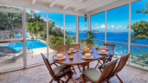 A covered outdoor dining area with a view of the sea, a pool, and a patio with lounge chairs.