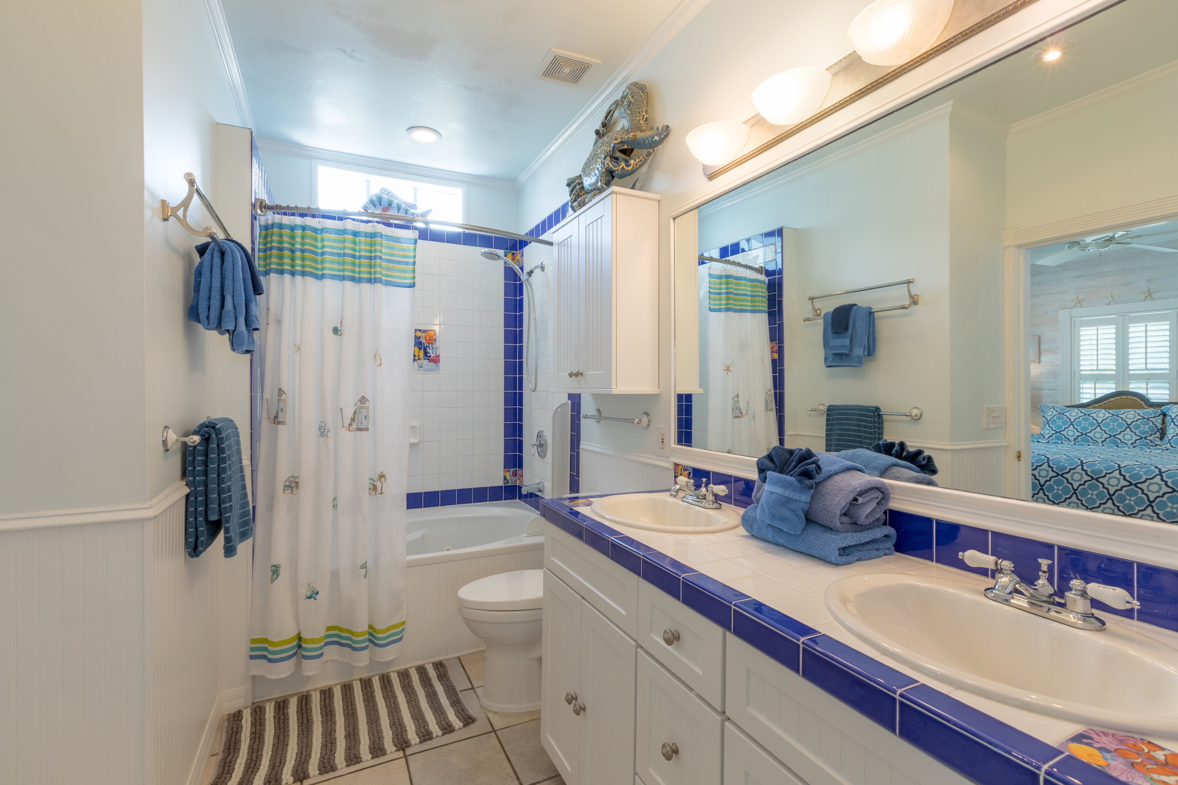 Un baño con bañera, inodoro y lavamanos, con azulejos azules y blancos, un espejo grande y una cortina de ducha a rayas.