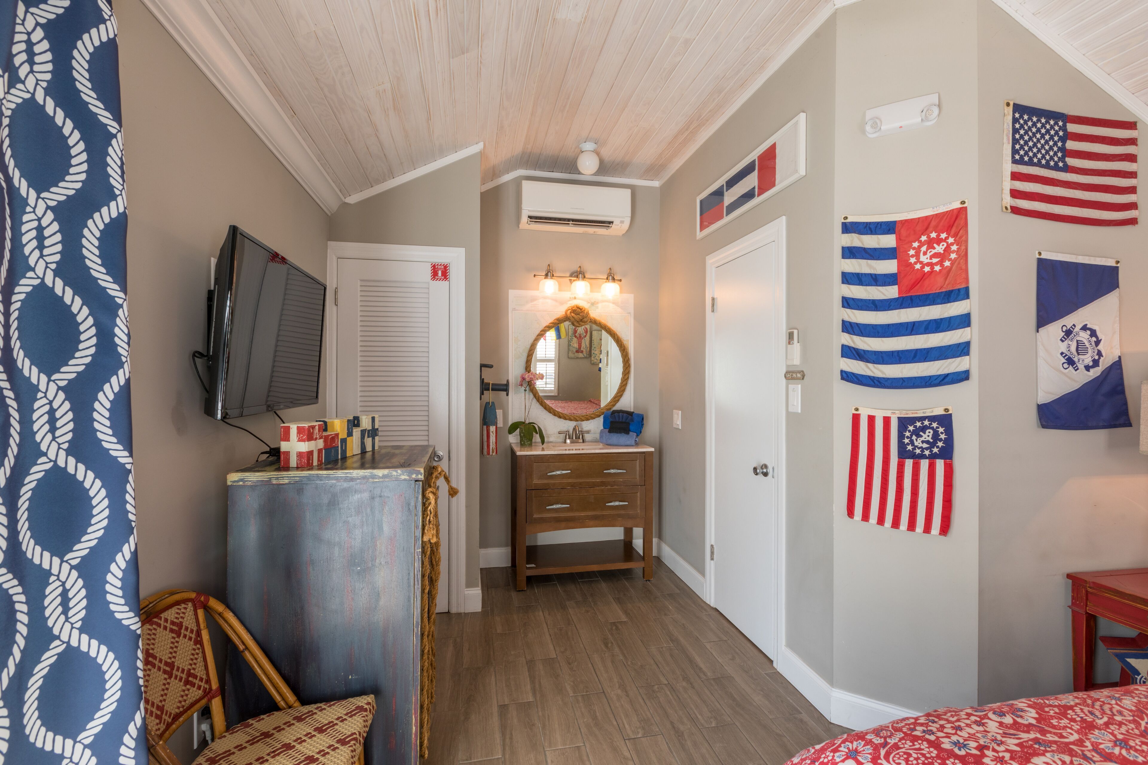 A bedroom with a wooden floor, a bed with a red and white patterned bedspread, a dresser, a mirror, and multiple flags on the wall.