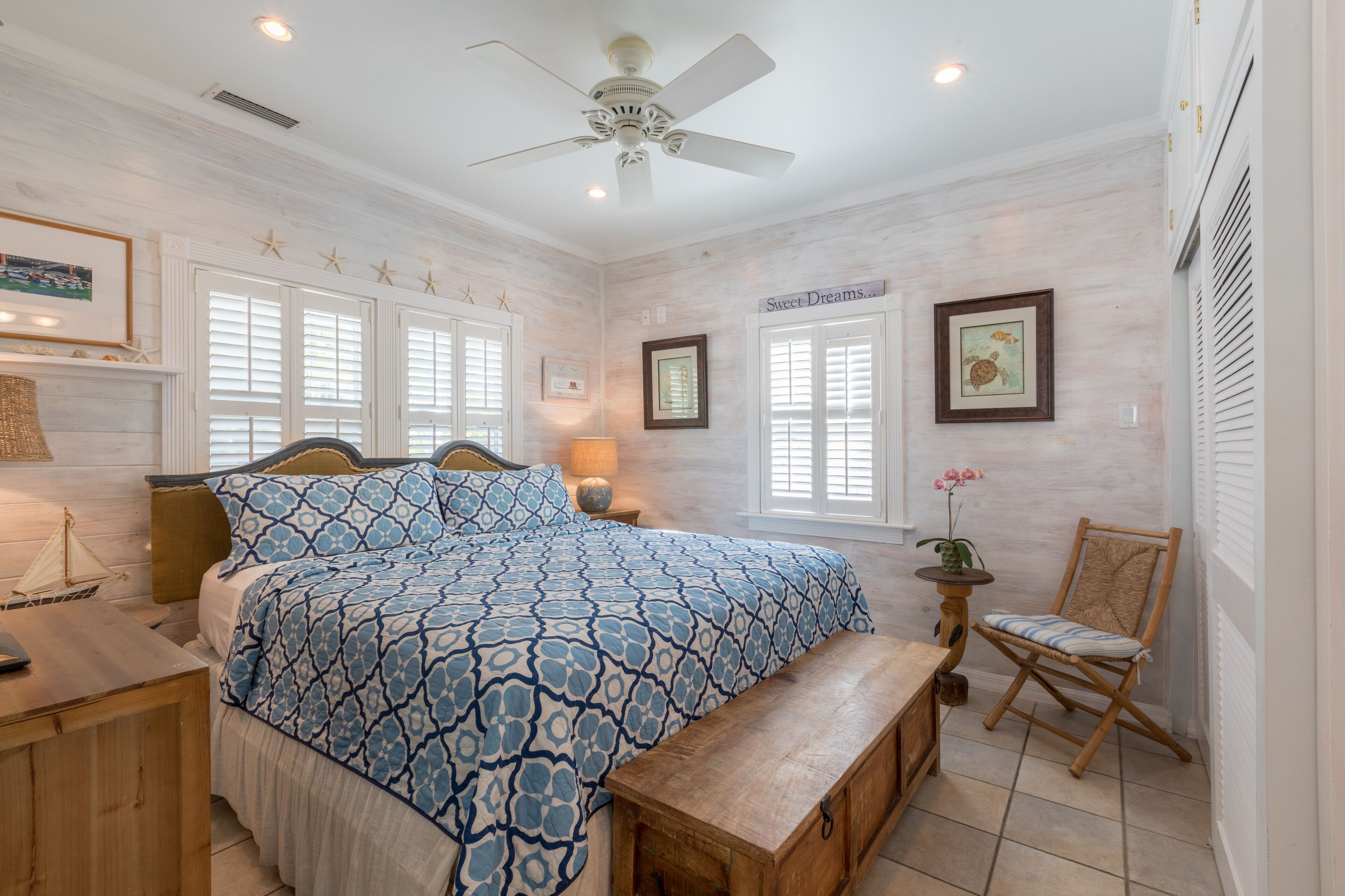 A bedroom with a large bed, a wooden bench, a chair, a ceiling fan, and a small table with a lamp.