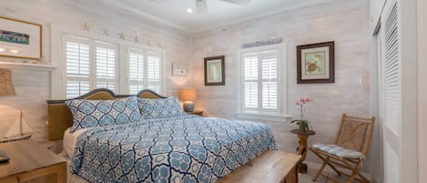 A bedroom with a large bed, a wooden bench, a chair, a ceiling fan, and a small table with a lamp.