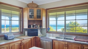 A kitchen with wooden cabinets, a built-in microwave, a toaster, and a dishwasher. There are large windows offering a view of the ocean. - 120 Ocean Parade Dalmeny (Dalmeny)