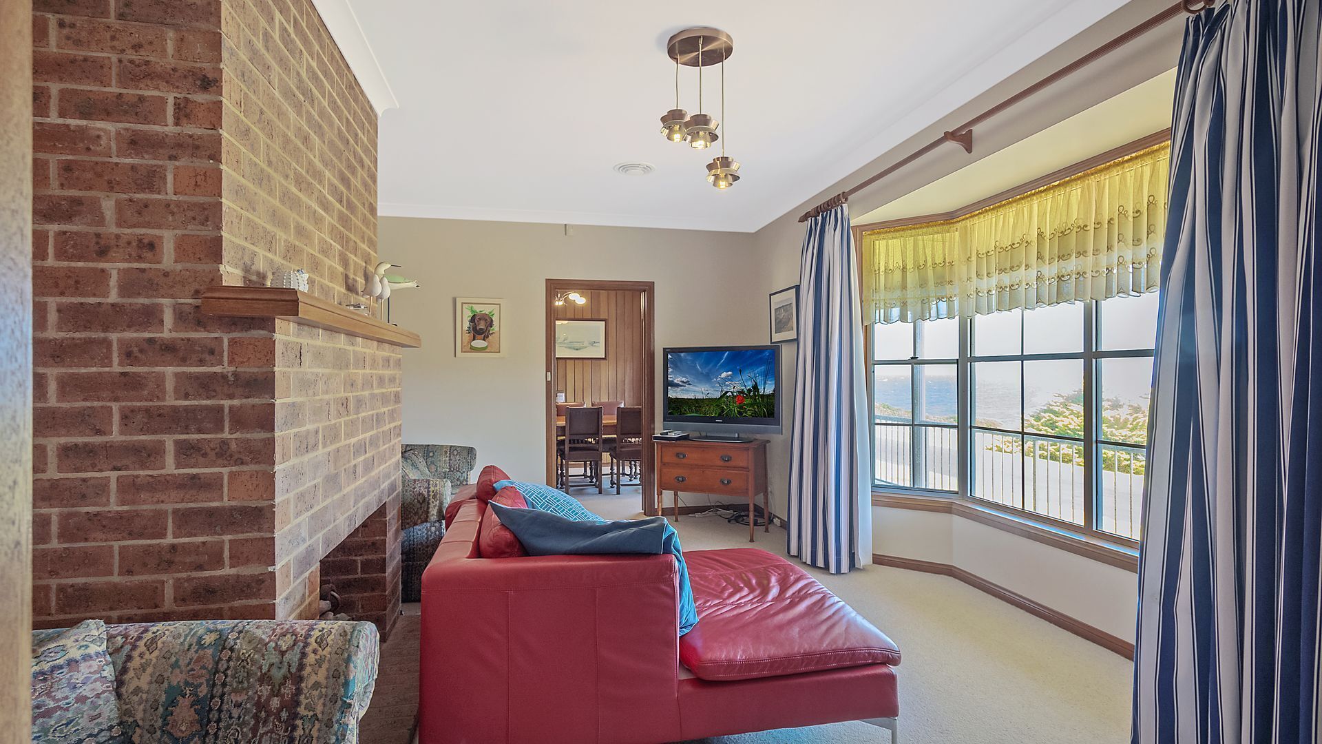 A room with a brick fireplace, a red sofa, a television, and a large window with curtains.