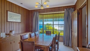 A dining room with a wooden table and chairs, a chandelier, and a view of the ocean through large windows. - 120 Ocean Parade Dalmeny (Dalmeny)