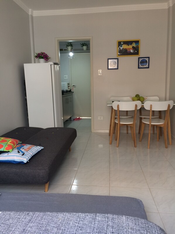 Un espace de vie compact comprenant un canapé, une table à manger avec des chaises, un réfrigérateur et un coin cuisine visible à travers une porte ouverte.