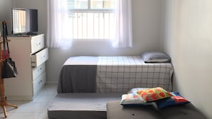 A small, well-lit bedroom with a bed, a desk with a computer, a window with curtains, and a radiator. - Cozy Kitnet At The Beach (Ponta da Praia)