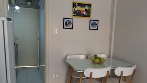 A dining area with a glass table and wooden chairs, a small fruit basket, and two framed pictures on the wall. - Cozy Kitnet At The Beach (Ponta da Praia)