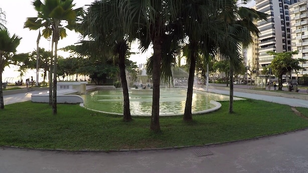 A circular water feature surrounded by palm trees and grass, with a paved walkway and a building in the background. - Cozy Kitnet At The Beach (Ponta da Praia)