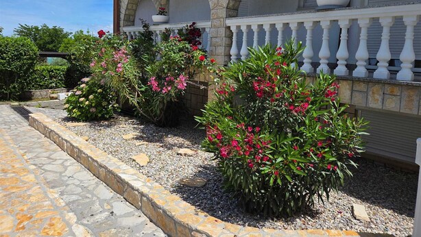 Un bâtiment à la façade en pierre, doté d’un balcon avec des balustrades blanches et d’un jardin agrémenté d’arbustes en fleurs.
