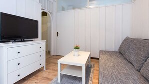 Un espace de vie compact avec une télévision posée sur une commode blanche, une petite table blanche ornée d’un vase et un canapé gris.