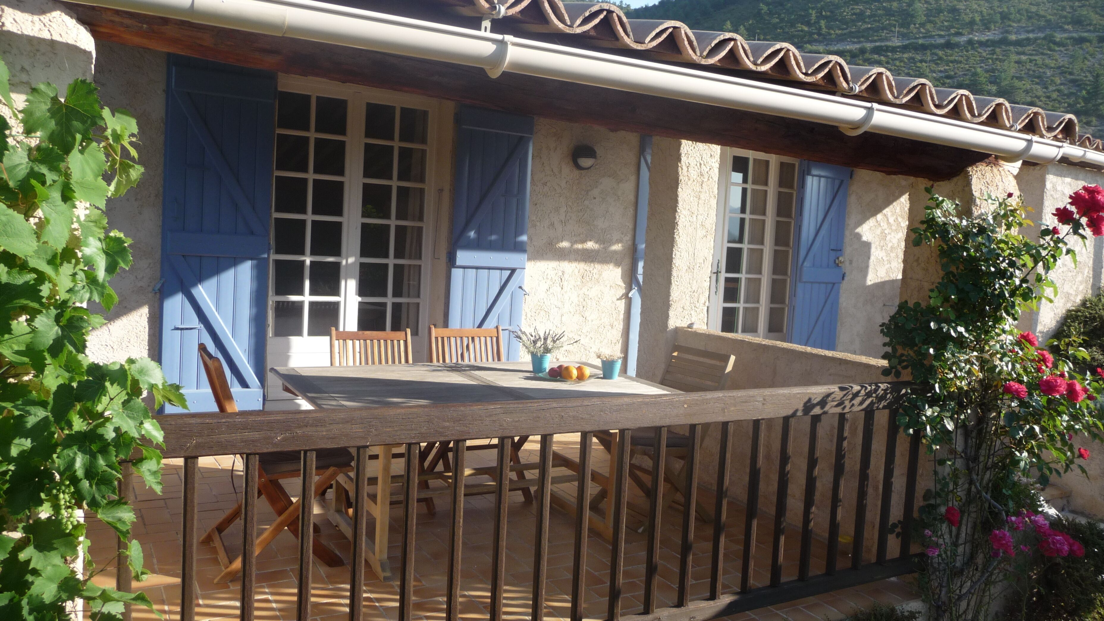 A rustic outdoor dining area with a wooden table and chairs, a blue door, and a stone wall.