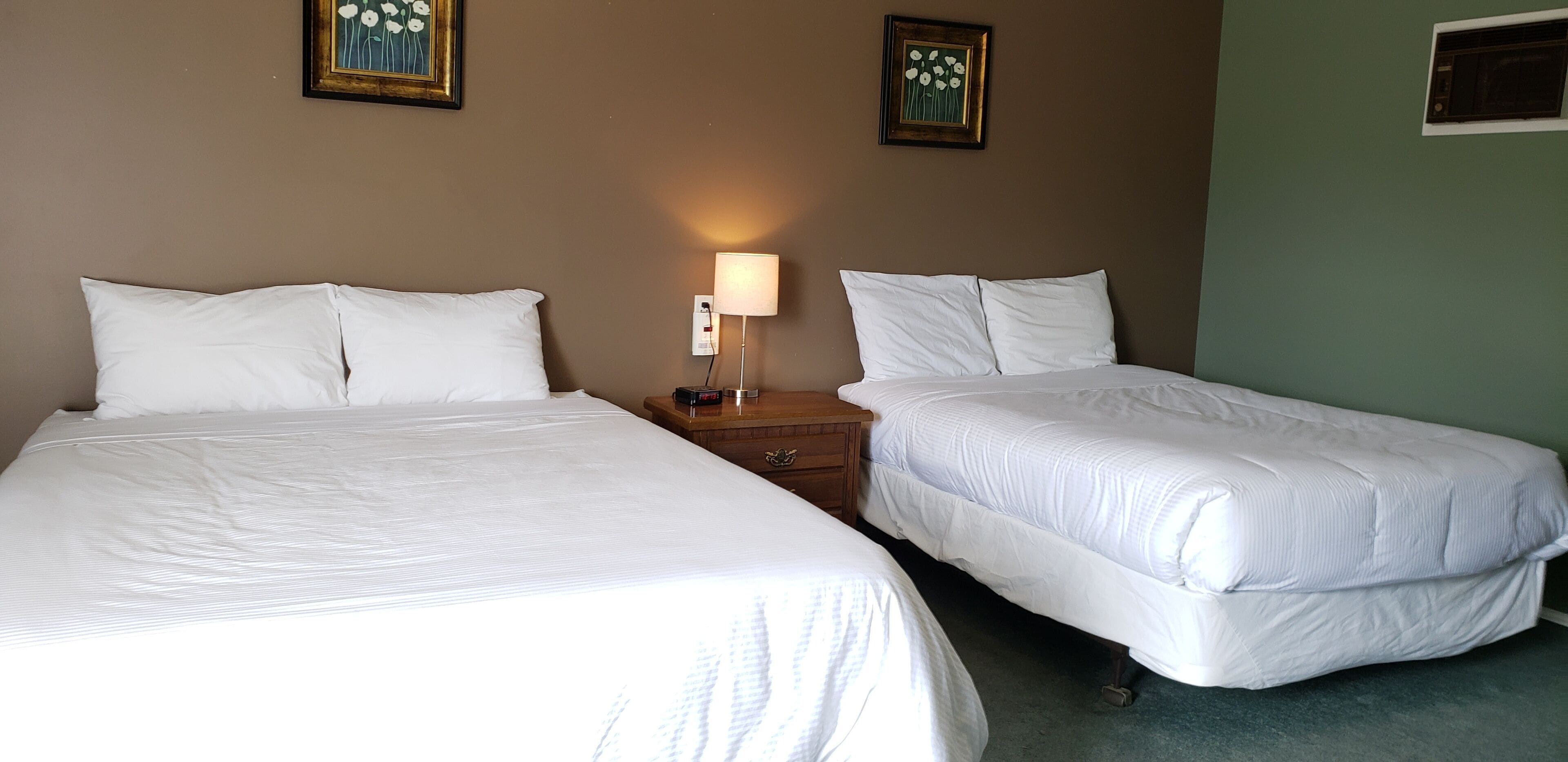 Una habitación de hotel con dos camas individuales, una mesita de noche con lámpara y dos cuadros enmarcados con motivos florales en la pared.