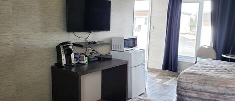 Habitación de hotel con televisor de pantalla plana en la pared, una kitchenette pequeña con microondas y cafetera, cama con cubrecama acolchado, y vista al exterior a través de una puerta de vidrio.