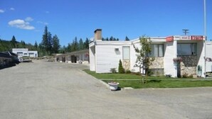 A row of single-story mobile homes with a gravel driveway, surrounded by trees and a clear sky.