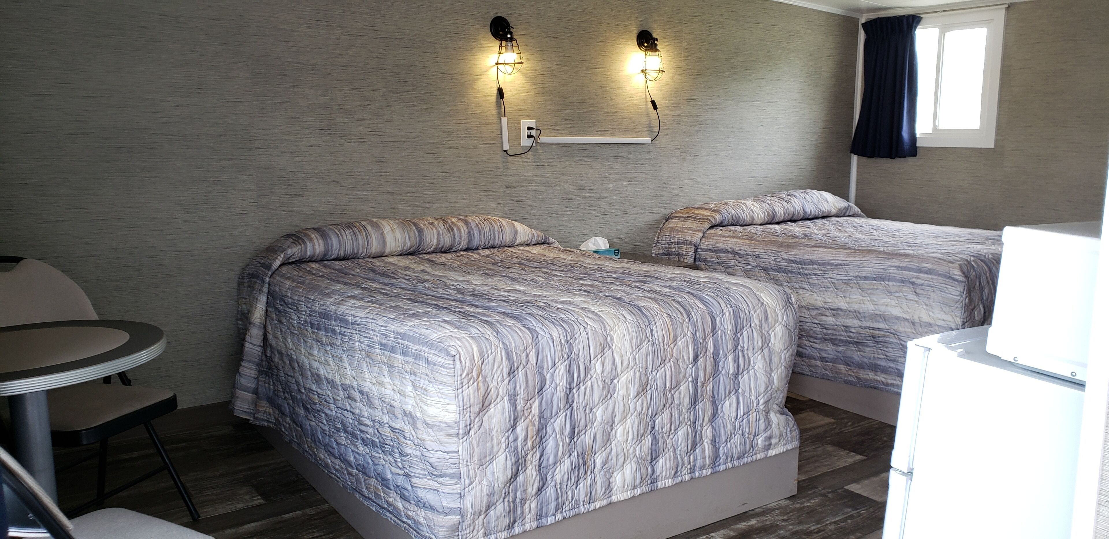 A hotel room with two beds, a small table, and a chair. There are wall-mounted lights and a window with curtains.